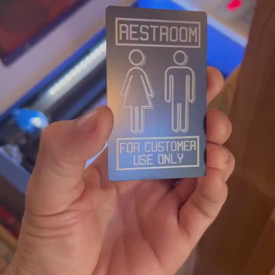 Close up video of hand holding a laser engraved metal retail signage placard, with Restroom and "For Customer Use Only" design