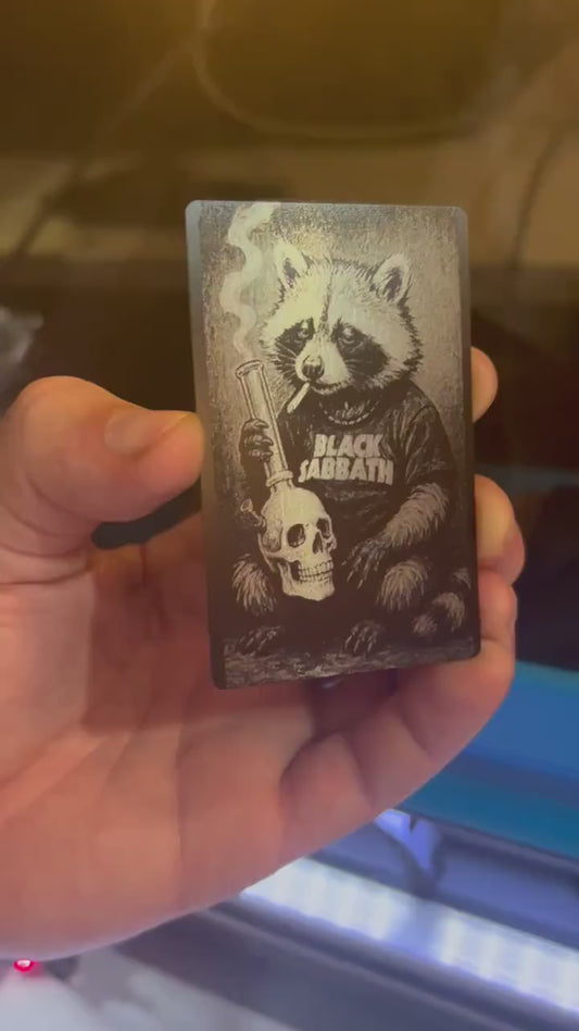 Hand holding a laser engraved metal/black illustrated cute animal gift card, with heavy metal raccoon design