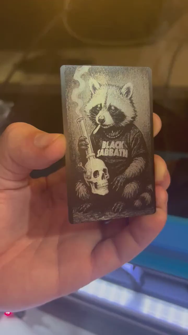Hand holding a laser engraved metal/black illustrated cute animal gift card, with heavy metal raccoon design