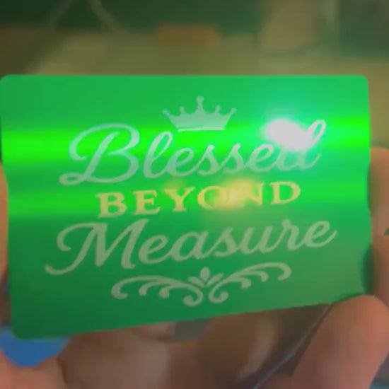 Hand holding a laser engraved metal green and silver anodized aluminum keepsake placard, with Blessed Beyond Measure typography with crown image in front of al laser engraving machine