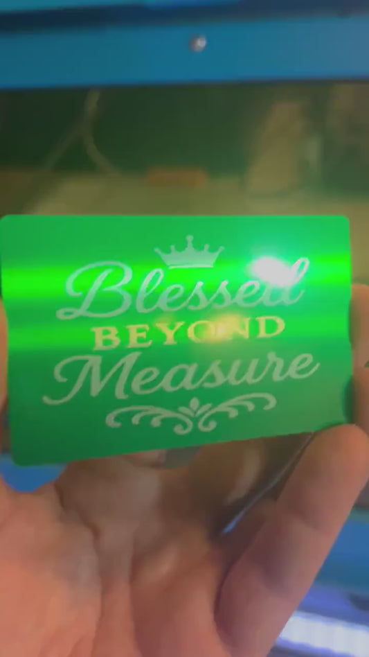 Hand holding a laser engraved metal green and silver anodized aluminum keepsake placard, with Blessed Beyond Measure typography with crown image in front of al laser engraving machine