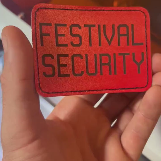 Hand holding a laser engraved red 3.5x2.5" Festival Security patch with heat activated adhesive backing in front of a laser engraving machine