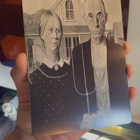 video close up of a hand holding a metal 5x7 laser engraved "American Gothic" plaque, in anodized aluminum rotating in the light to demonstrate reflective qualities