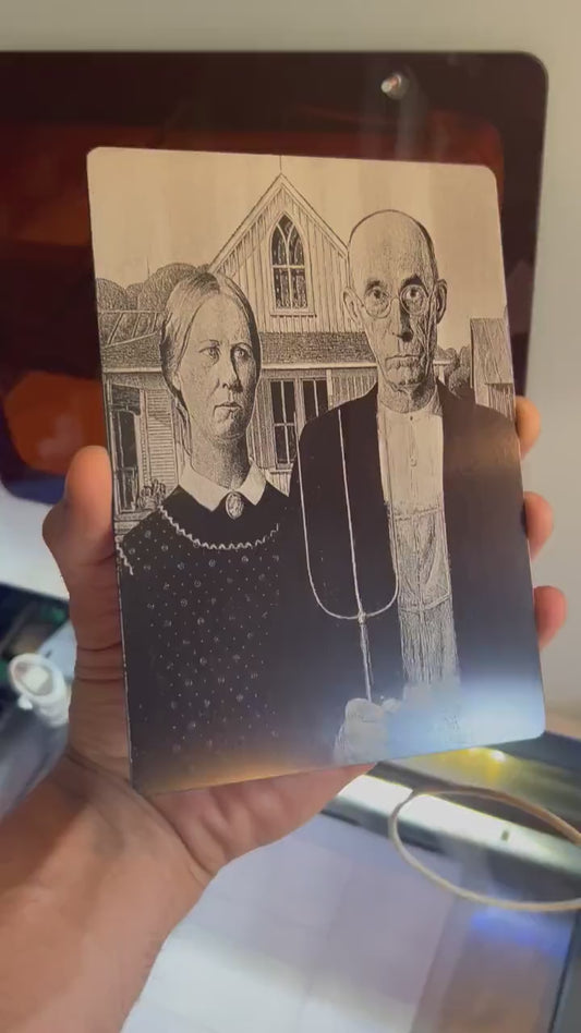 video close up of a hand holding a metal 5x7 laser engraved "American Gothic" plaque, in anodized aluminum rotating in the light to demonstrate reflective qualities