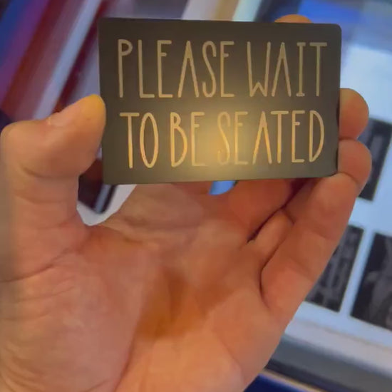 Video of engraved metal restaurant placard with Please Wait To Be Seated silver text, turning under light to show reflective finish