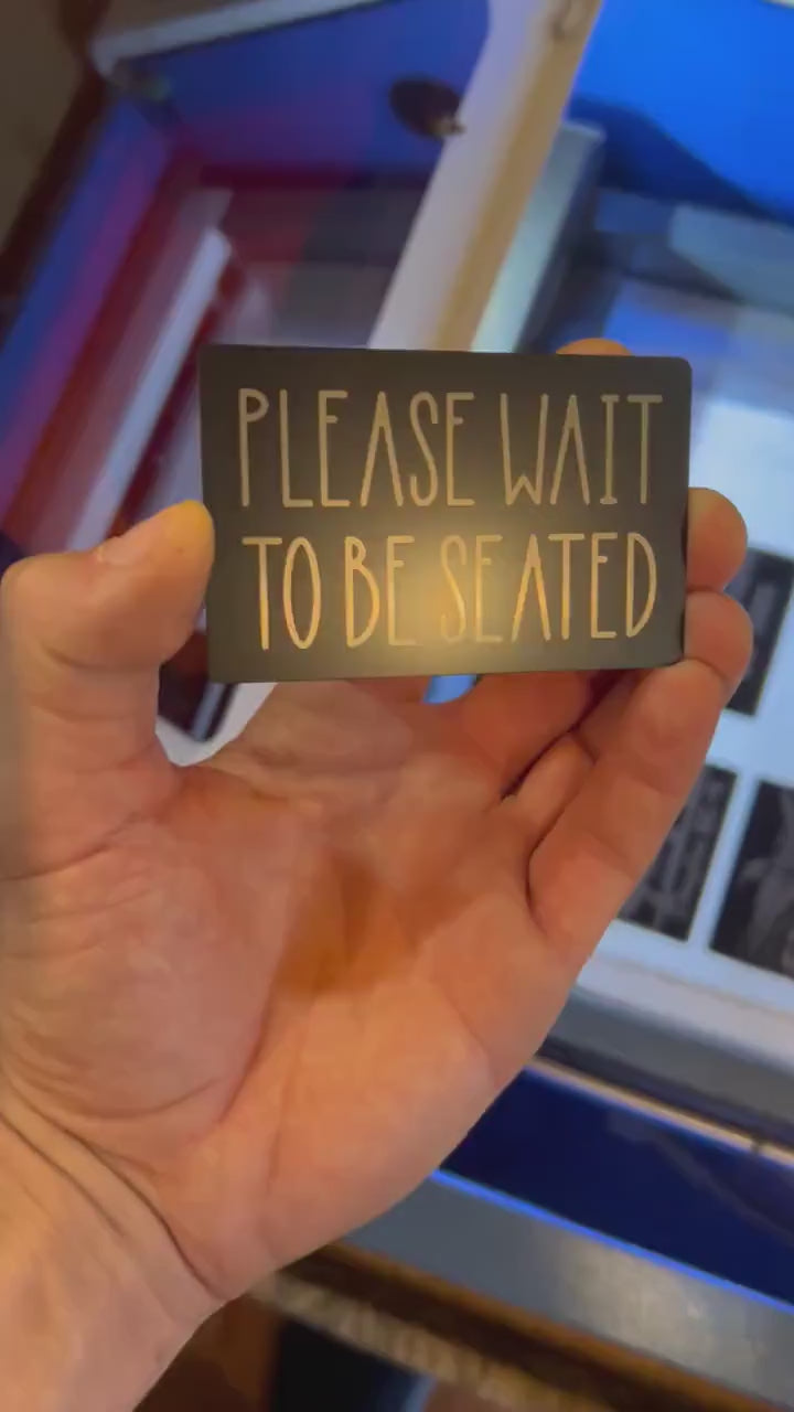 Video of engraved metal restaurant placard with Please Wait To Be Seated silver text, turning under light to show reflective finish