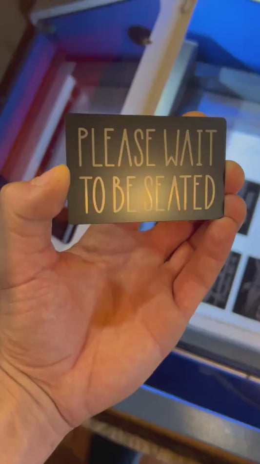Video of engraved metal restaurant placard with Please Wait To Be Seated silver text, turning under light to show reflective finish