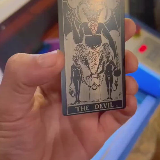 Video of hand holding a black/silver metal tarot card with The Devil design and text