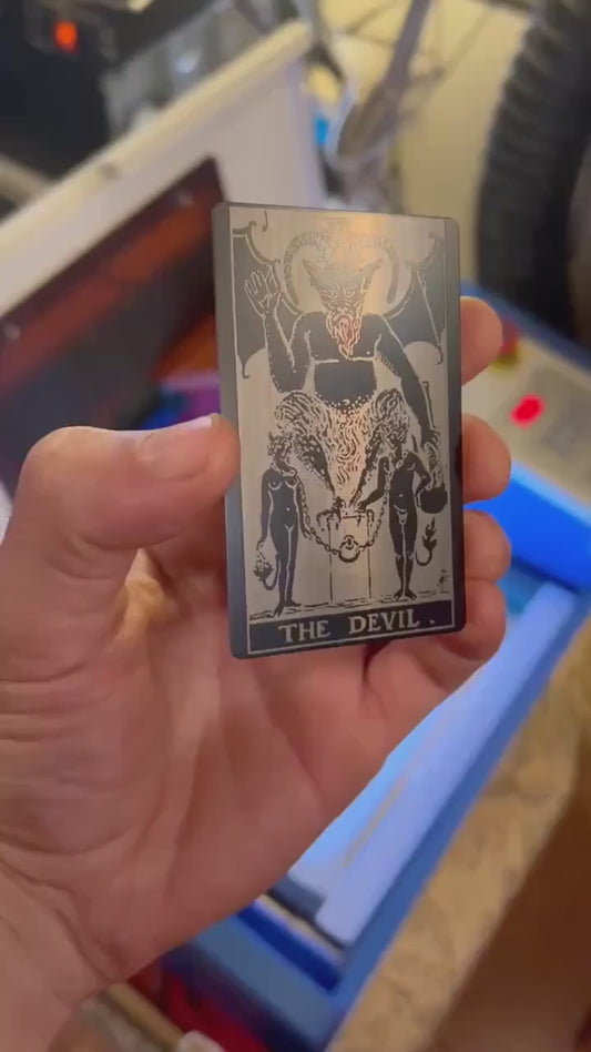 Video of hand holding a black/silver metal tarot card with The Devil design and text