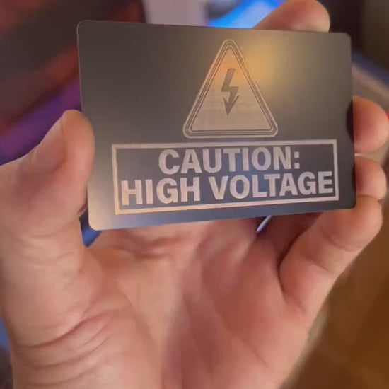 Close up video of hand holding a laser engraved metal safety warning signage, with Caution High Voltage and an electrical symbol