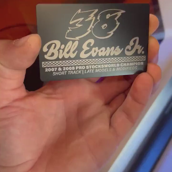 Hand holding a metal black/silver anodized aluminum metal race car driver's business card for marketing and promotional events, demonstrated in front of a laser engraving machine