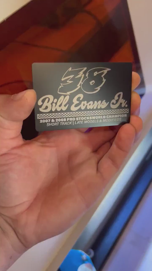 Hand holding a metal black/silver anodized aluminum metal race car driver's business card for marketing and promotional events, demonstrated in front of a laser engraving machine