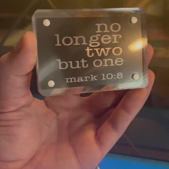 Hand holding a laser engraved metal Christian gift card that reads "No Longer Two, But One" with bible verse reference for Mark 10:8 in black/silver anodized aluminum