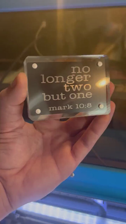 Hand holding a laser engraved metal Christian gift card that reads "No Longer Two, But One" with bible verse reference for Mark 10:8 in black/silver anodized aluminum