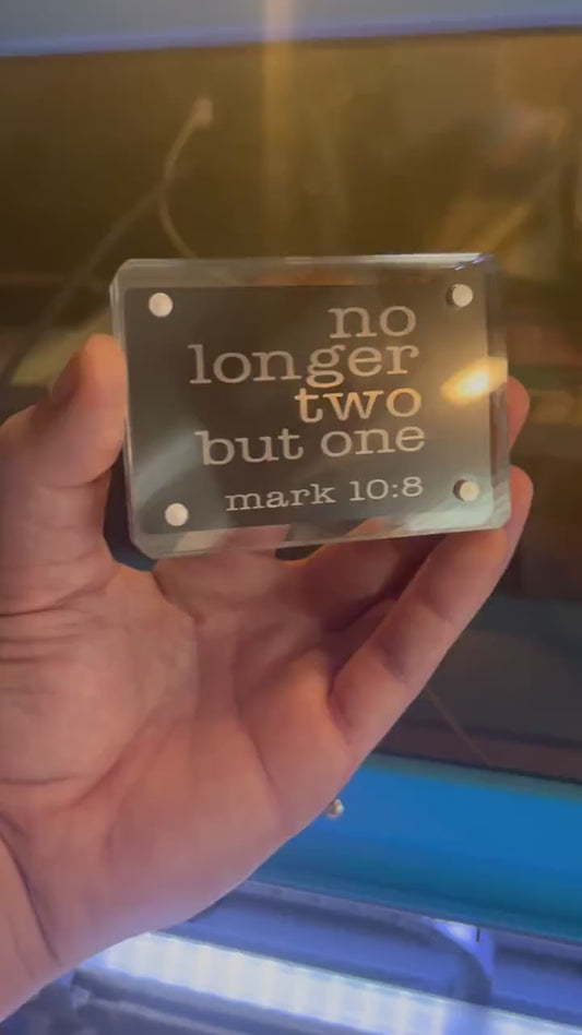 Hand holding a laser engraved metal Christian gift card that reads "No Longer Two, But One" with bible verse reference for Mark 10:8 in black/silver anodized aluminum