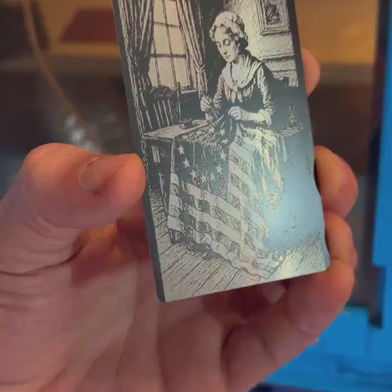 Close up video of hand holding a laser engraved metal inspirational keepsake card with Betsy Ross sewing the first American flag illustration artwork in black/silver
