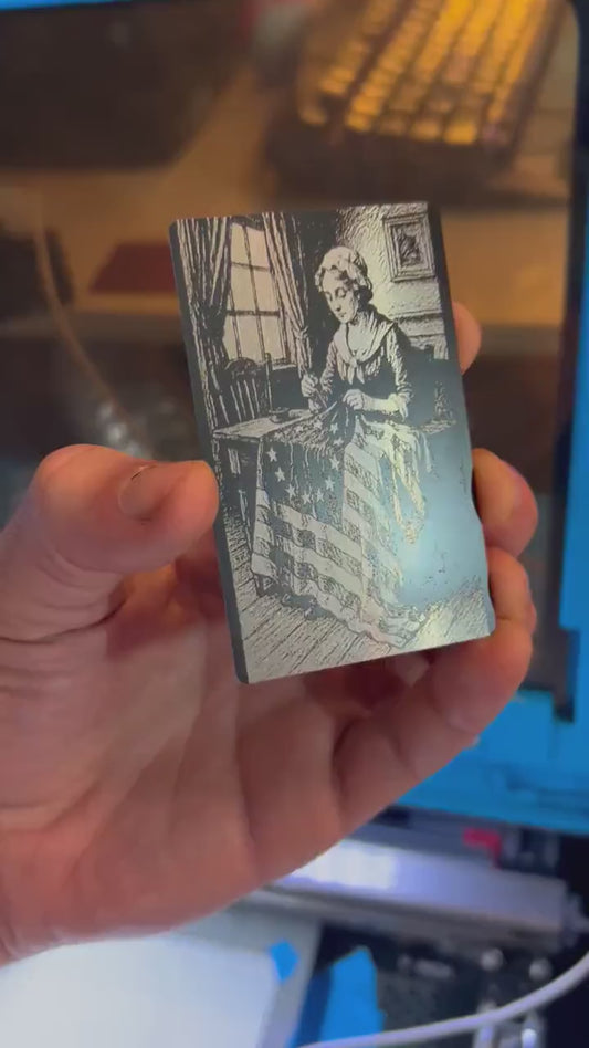 Close up video of hand holding a laser engraved metal inspirational keepsake card with Betsy Ross sewing the first American flag illustration artwork in black/silver