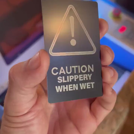 hand holding a metal retail signage placard with caution slippery when wet keepsake funny gift card in front of a laser engraving machine