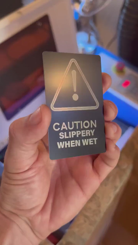 hand holding a metal retail signage placard with caution slippery when wet keepsake funny gift card in front of a laser engraving machine