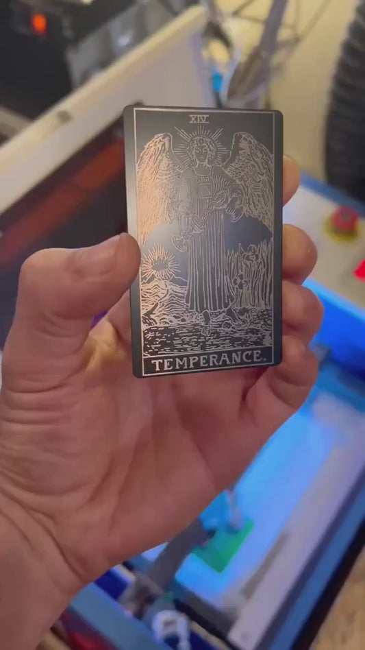 Video of hand demonstrating a black/silver metal Temperance tarot card design under lighting showing how reflective the metal is
