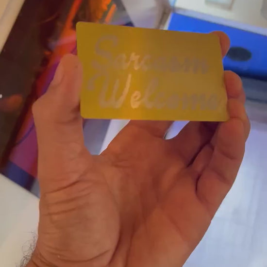 Hand holding a gold over silver laser engraved metal business card, with novelty Sarcasm Welcome customized engraving, in front of a laser engraving machine