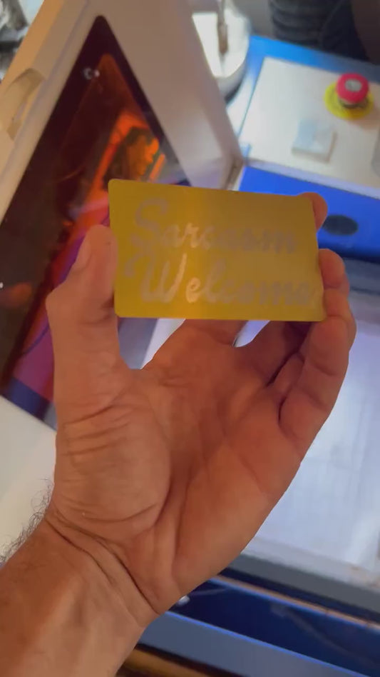 Hand holding a gold over silver laser engraved metal business card, with novelty Sarcasm Welcome customized engraving, in front of a laser engraving machine