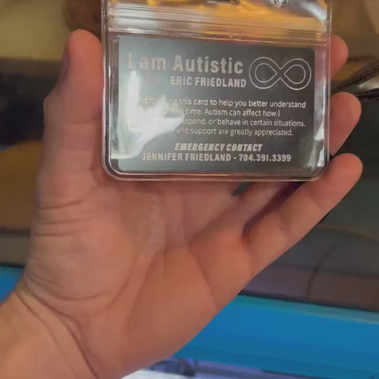 Hand demonstrating a laser engraved metal business card with black/silver design shown in a plastic badge holder with lanyard in front of a cnc machine