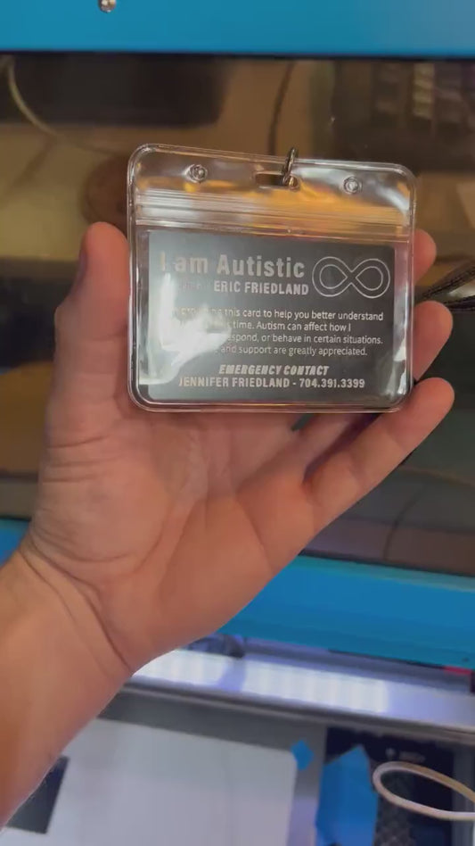 Hand demonstrating a laser engraved metal business card with black/silver design shown in a plastic badge holder with lanyard in front of a cnc machine