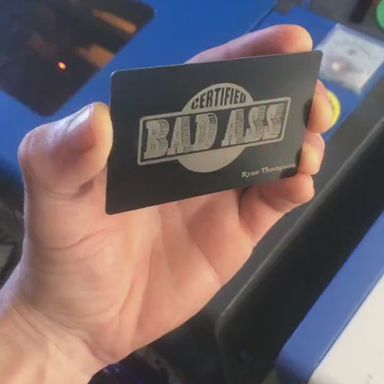 Close up video of hand holding a laser engraved metal gift card with Certified Bad Ass Text, and custom personalized name below