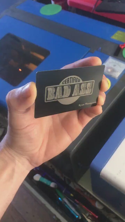 Close up video of hand holding a laser engraved metal gift card with Certified Bad Ass Text, and custom personalized name below