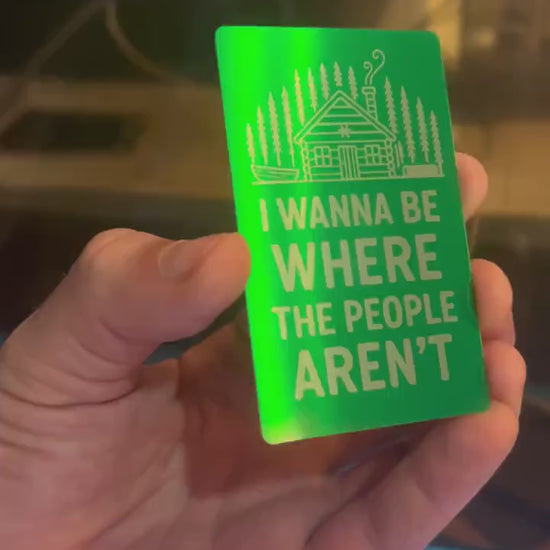 Hand holding a laser engraved metal keepsake card with "I Wanna Be Where The People Aren't" text and cabin graphics, demonstrating reflective qualities of card under light