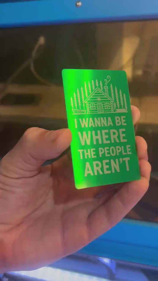 Hand holding a laser engraved metal keepsake card with "I Wanna Be Where The People Aren't" text and cabin graphics, demonstrating reflective qualities of card under light