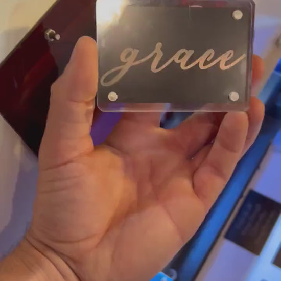 Hand holding a laser engraved metal keepsake card that reads "Grace" in stylish cursive handwritten typography