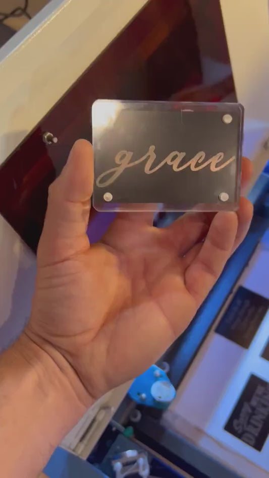 Hand holding a laser engraved metal keepsake card that reads "Grace" in stylish cursive handwritten typography