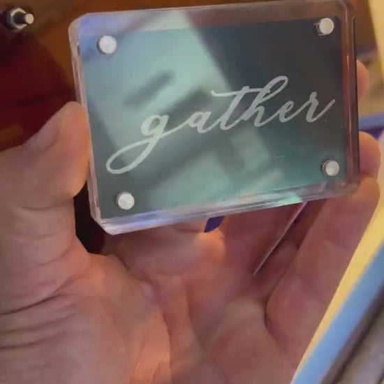 Detailed shot of anodized aluminum mini keepsake placard in acrylic display case, engraved with “gather” text