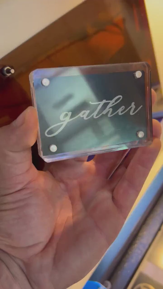 Detailed shot of anodized aluminum mini keepsake placard in acrylic display case, engraved with “gather” text