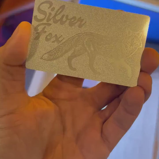 Showcasing a pearl silver and silver metal friendsgiving gift card that reads "Silver Fox" with fox graphic in front of blurred machine background