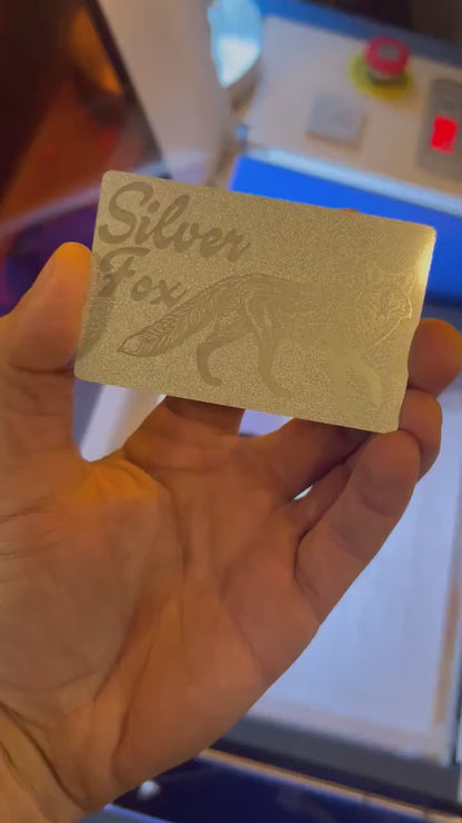 Showcasing a pearl silver and silver metal friendsgiving gift card that reads "Silver Fox" with fox graphic in front of blurred machine background