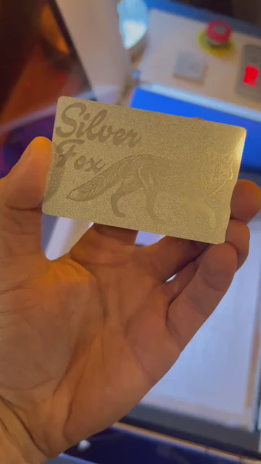 Showcasing a pearl silver and silver metal friendsgiving gift card that reads "Silver Fox" with fox graphic in front of blurred machine background