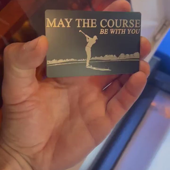 video of hand holding and rotating a laser engraved metal keepsake card with "May the course be with you" engraved text and golfer making golf swing on beautiful golf course art