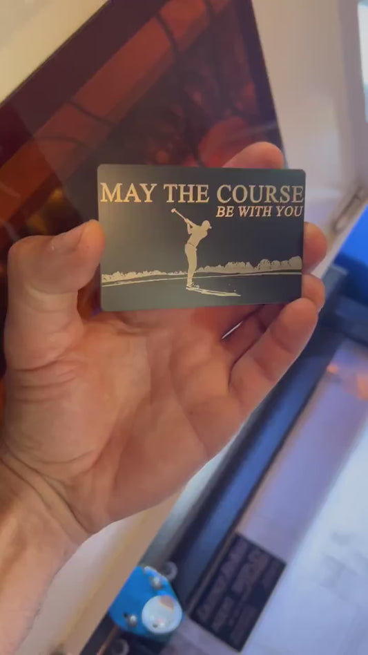 video of hand holding and rotating a laser engraved metal keepsake card with "May the course be with you" engraved text and golfer making golf swing on beautiful golf course art