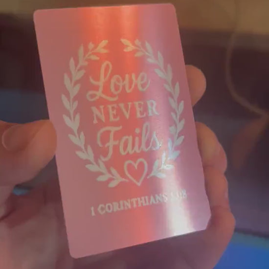 Close-up of hand displaying a rose gold metal keepsake card with “Love Never Fails” typography design and 1 Corinthians 13:8 scripture