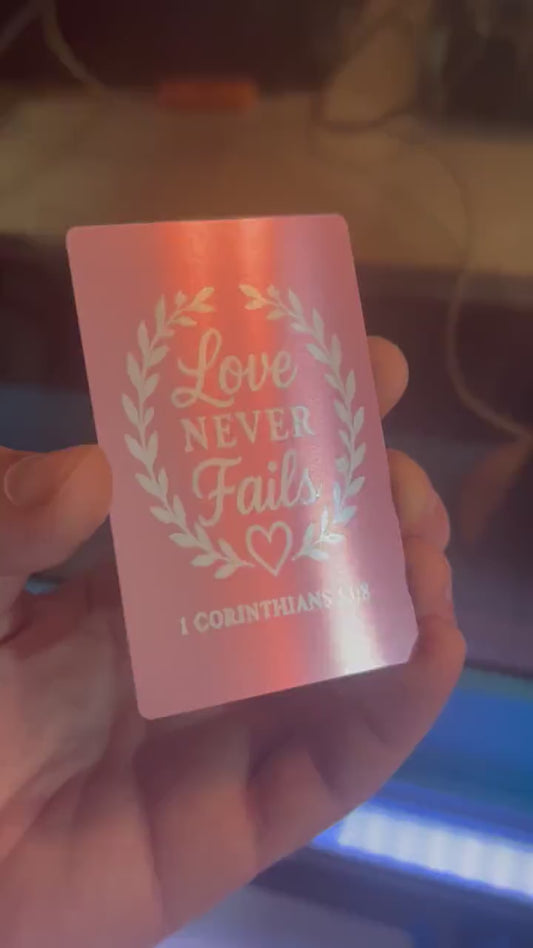 Close-up of hand displaying a rose gold metal keepsake card with “Love Never Fails” typography design and 1 Corinthians 13:8 scripture