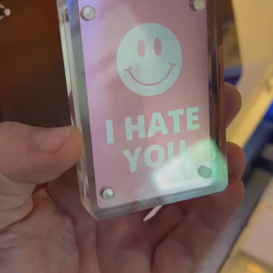 Hand holding metal I Hate You with smiley face graphic laser engraving card, shown in optional Acrylic Display Case
