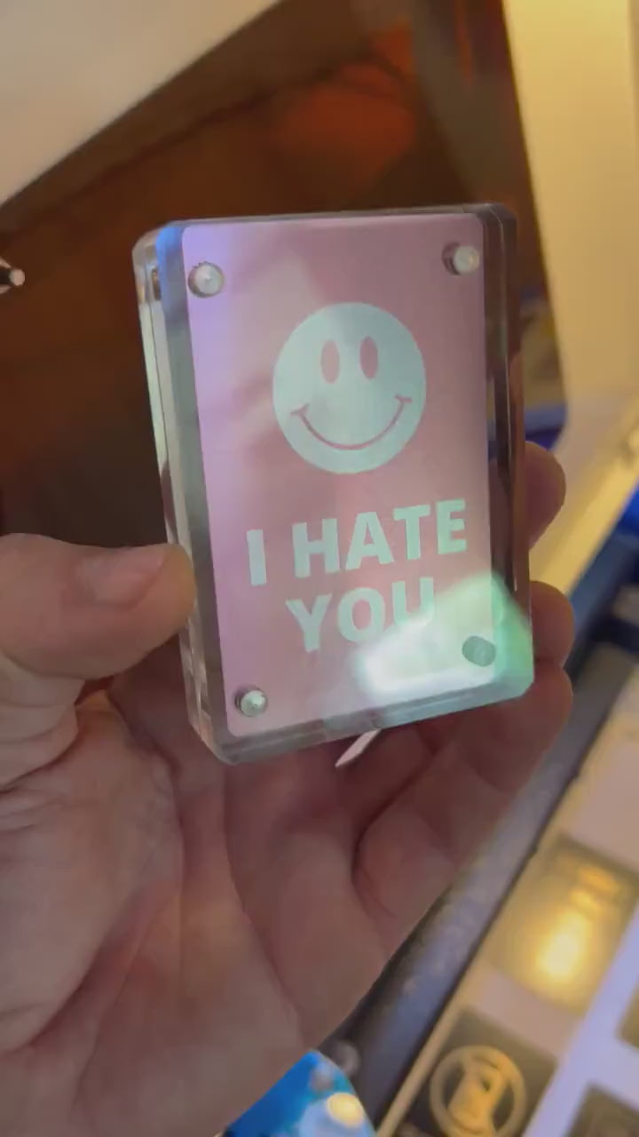 Hand holding metal I Hate You with smiley face graphic laser engraving card, shown in optional Acrylic Display Case