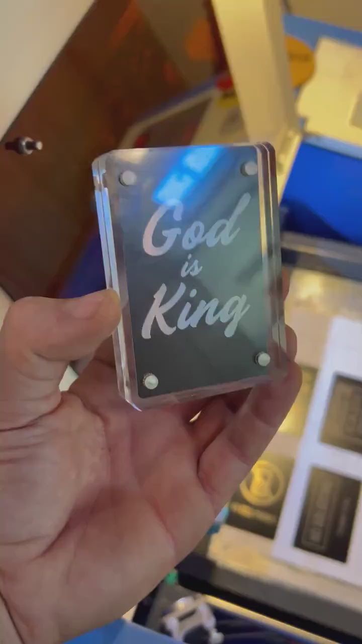 Hand holding a laser engraved black/silver God is King metal bookmark card, shown in optional acrylic display case