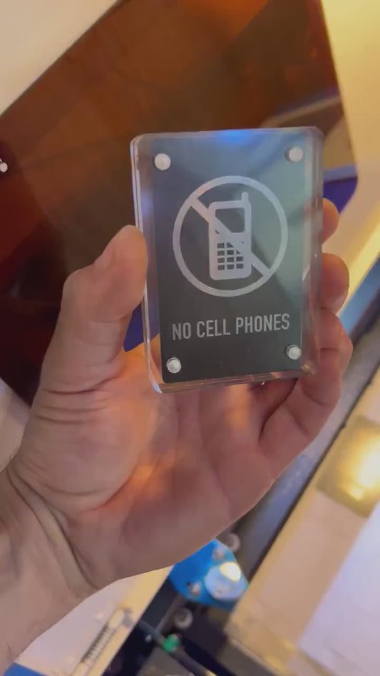Hand holding a laser engraved black/silver No Cell Phones metal signage shown in optional acrylic display case