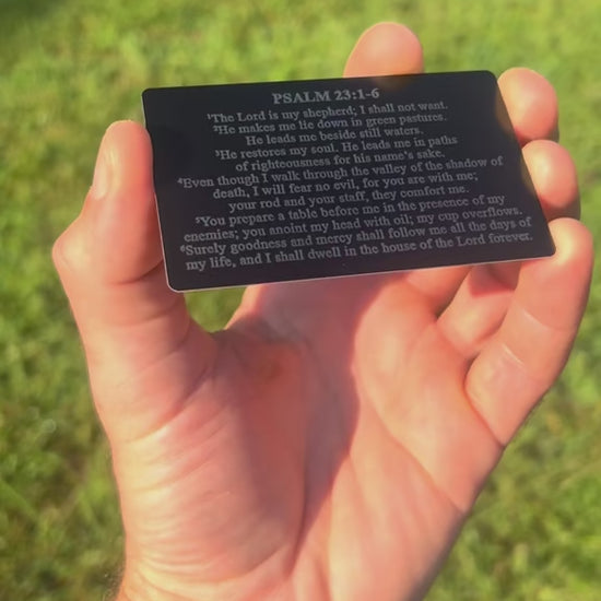 Hand holding a laser engraved metal bible verse card in front of a natural background