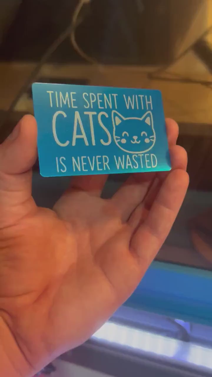 Hand holding a laser engraved metal pet lover gift that reads "Time Spent With Cats Is Never Wasted", with cute kitten graphic in blue/silver anodized aluminum