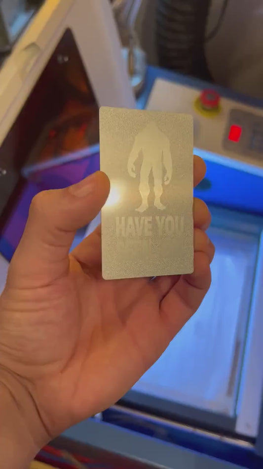 Close up video of hand holding a laser engraved metal gift card with Have you seen him? text and bigfoot silhouette artwork design in SIlver/Silver